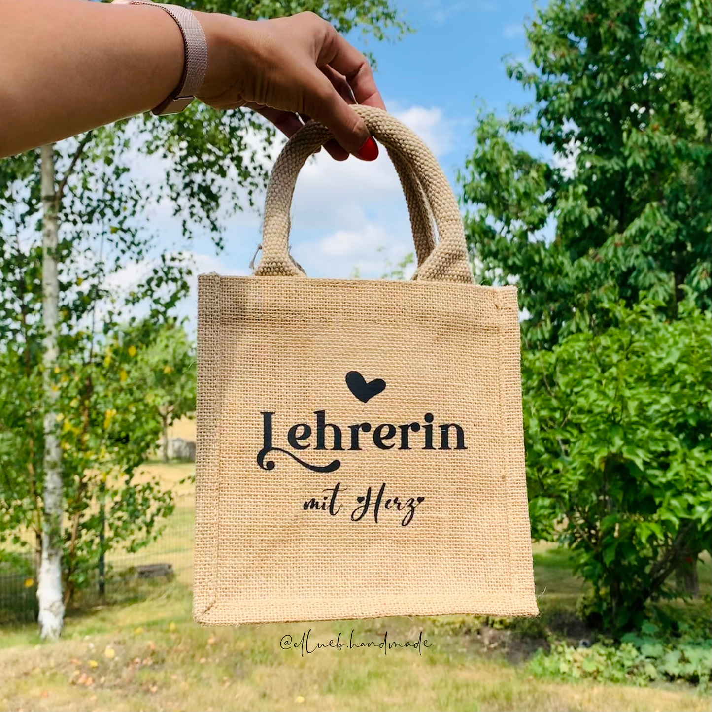 Ein hand hält die kleine Jutetasche hoch. Im Hintergrund sind grüne Bäume und blauer Himmel. Auf dem Jutetasche steht Lehrerin mit Herz mit ein kleiner Herz.