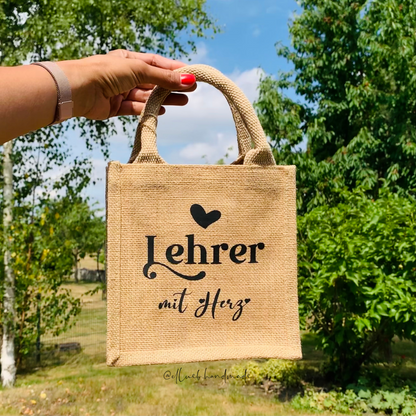 Ein hand hält die kleine Jutetasche hoch. Im Hintergrund sind grüne Bäume und blauer Himmel. Auf dem Jutetasche steht Lehrer mit Herz mit ein kleiner Herz. 