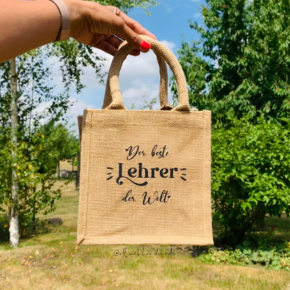 Ein hand hält die kleine Jutetasche hoch. Im Hintergrund sind grüne Bäume und blauer Himmel. Auf dem Jutetasche steht ,,Die beste Lehrer der Welt".  