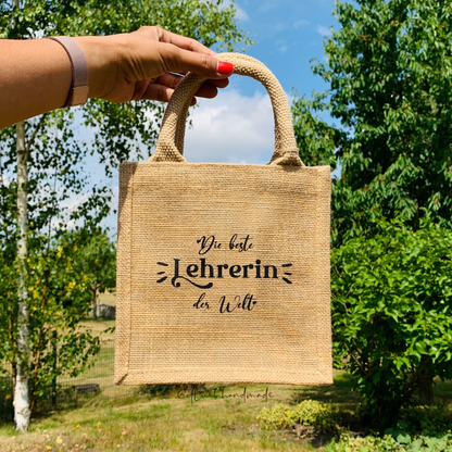 Ein hand hält die kleine Jutetasche hoch. Im Hintergrund sind grüne Bäume und blauer Himmel.