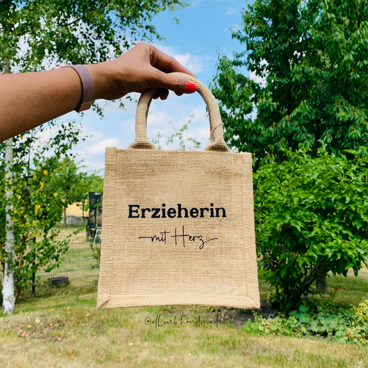 Ein hand hält die kleine Jutetasche hoch. Im Hintergrund sind grüne Bäume und blauer Himmel. Auf dem Jutetasche steht Erzieherin mit Herz mit ein kleiner Herz.